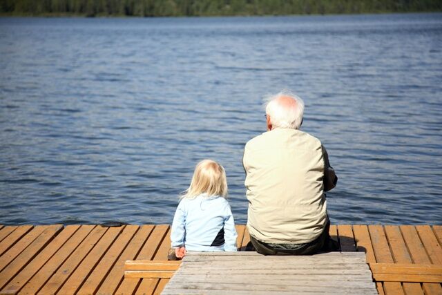 Großvater und blondes Enkelkind sitzen auf einem Steg am Wasser. Rückenansicht