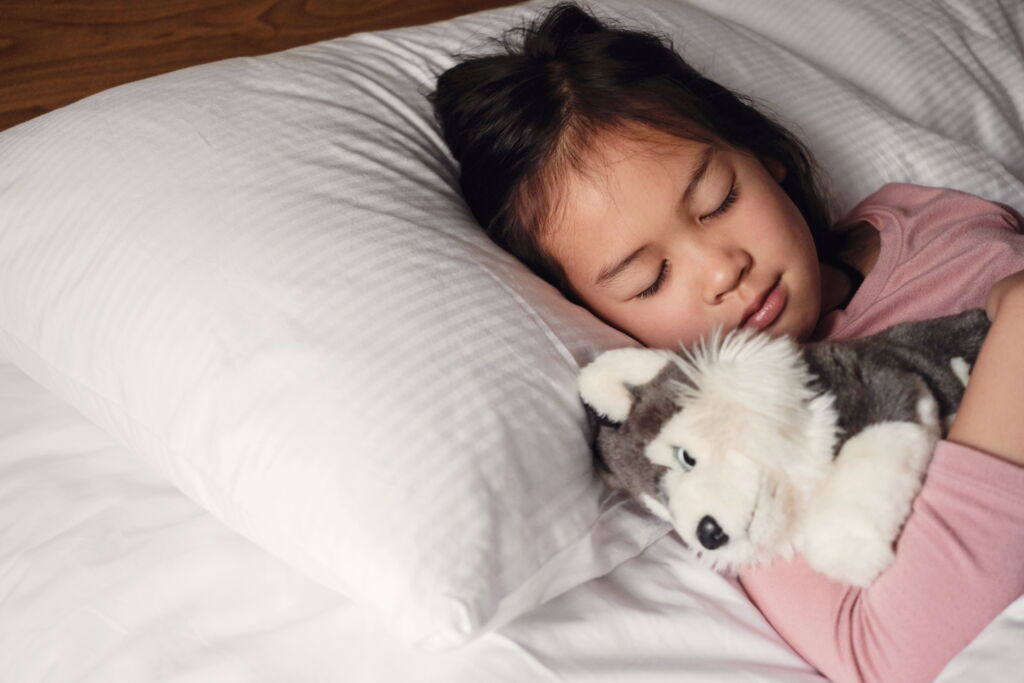 dunkelhaariges, schlafendes Mädchen mit Kuscheltier in Rückenlage, weiße Bettwäsche