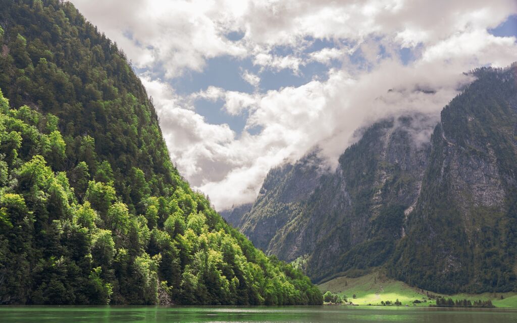 Landschaft. Grüner See. Steile Berge, bis zur Wasserlinie mit grünen Bäumen bewachsen, locker bewölkter Himmel